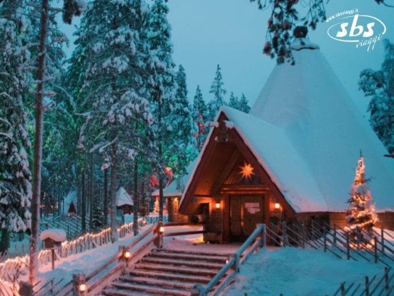 Una pittoresca scena di baita innevata a Rovaniemi, con un sentiero illuminato e un albero di Natale incastonato in un ambiente boschivo. L'edificio con tetto a punta si trova sotto un cielo che si oscura, creando il perfetto paese delle meraviglie invernale nordico.