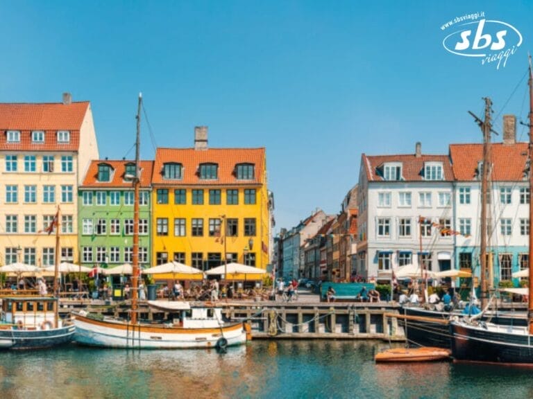 Edifici colorati costeggiano il lungomare di Copenaghen, con barche attraccate di fronte sotto un cielo limpido e azzurro.