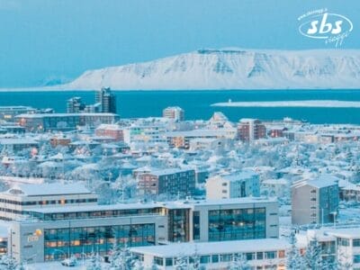 Un paesaggio urbano innevato in Islanda, con edifici e strade ricoperti di neve. Montagne e mare sono visibili sullo sfondo, mentre l'aurora boreale danza nel cielo, creando una magica atmosfera di Capodanno.