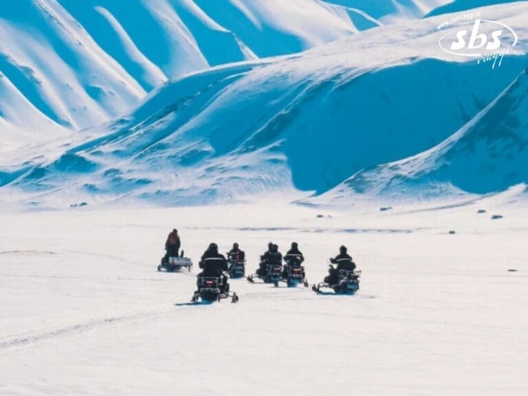 Un gruppo di persone su motoslitte attraversa un vasto paesaggio innevato, intraprendendo avventure tra i ghiacci con le maestose montagne dell'Islanda sullo sfondo.