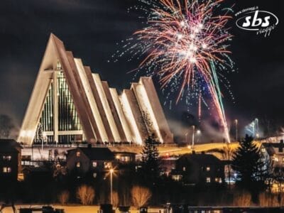 I fuochi d'artificio illuminano il cielo notturno sopra un moderno edificio angolare con grandi finestre triangolari, che ricordano il fascino di Tromso, circondato da palazzi e lampioni. La scena sembra una celebrazione del Capodanno artico, con colori vivaci che danzano come un'aurora boreale in cielo.