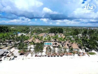 Vista aerea dello Sbh Monica Zanzibar, uno splendido resort sulla spiaggia caratterizzato da edifici con tetti di paglia incastonati tra palme ondeggianti, con una tranquilla piscina al centro e un cielo nuvoloso che proietta dolci ombre sopra.