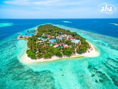 Vista aerea di una piccola e lussureggiante isola circondata da limpide acque turchesi, con l'elegante Plumeria Hotel tra vari edifici e una spiaggia sabbiosa.