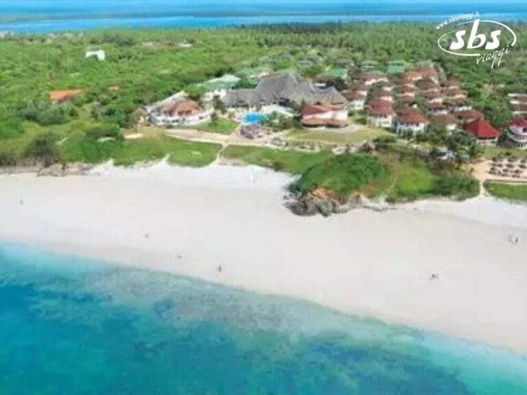 Vista aerea di un resort sulla spiaggia con sabbia bianca, mare azzurro e vegetazione verde. Numerosi edifici e una piscina sono visibili all'interno del complesso del resort.