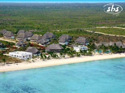 Vista aerea di un resort costiero con edifici dal tetto di paglia, una spiaggia di sabbia bianca e acque cristalline. Una fitta vegetazione verde circonda il resort Bozza Automatica, offrendo un'armoniosa miscela di natura e tranquillità.