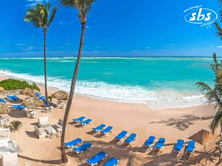 Una scena di spiaggia a Whala! Bavaro presenta acqua turchese, palme e file di sedie a sdraio blu su sabbia bianca sotto un cielo azzurro e terso.