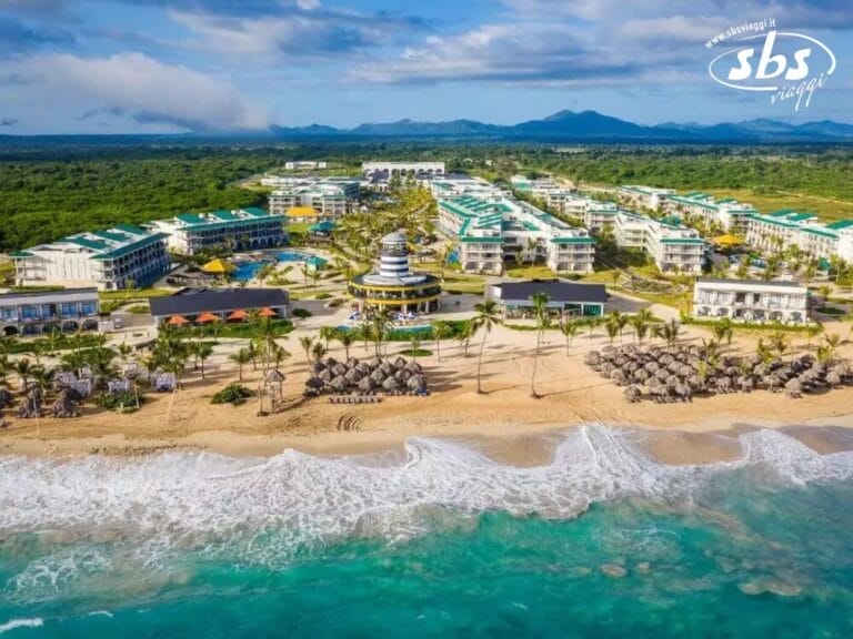 Vista aerea di un resort sulla spiaggia con edifici bianchi, palme e ombrelloni di paglia che costeggiano la riva sabbiosa. Le onde dell'oceano blu raggiungono dolcemente la spiaggia, creando una serena bozza automatica che cattura perfettamente il paradiso.