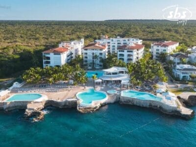 Vista aerea di un resort costiero con più edifici, circondato da alberi rigogliosi. Due grandi piscine, come splendidi schizzi di bozza automatica, sono visibili vicino alla costa rocciosa, con lettini disposti ad arte intorno a loro.