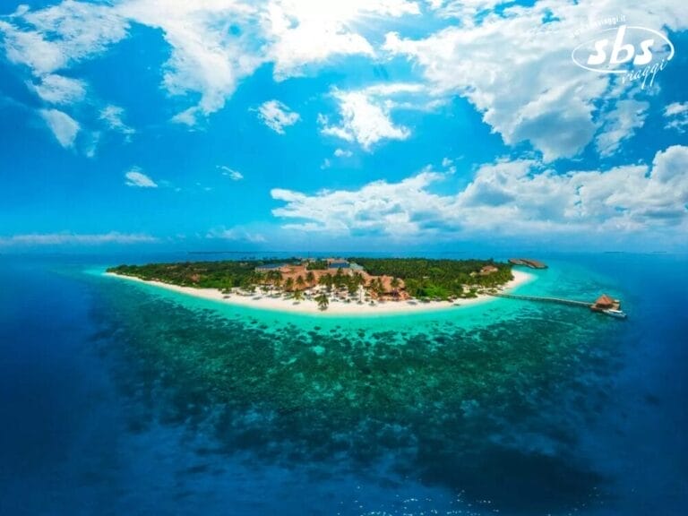 Vista aerea del Reethi Faru Resort su un'isola tropicale circondata dalle acque blu dell'oceano, con un molo che si estende dalla riva e nuvole sparse in un cielo luminoso.