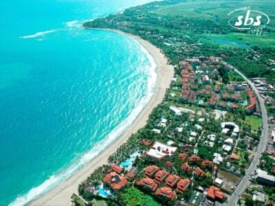 Da una prospettiva aerea, la zona costiera rivela una straordinaria miscela di lunghe spiagge sabbiose, acque turchesi e vegetazione lussureggiante. Edifici con tetti rossi punteggiano il paesaggio mentre le strade serpeggiano lungo la costa, creando una pittoresca bozza automatica di bellezza naturale e tocco umano.