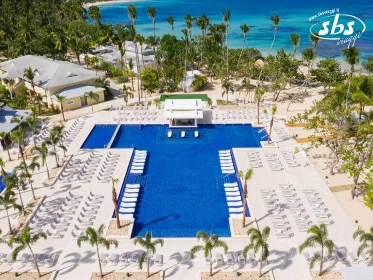 Vista aerea del resort tropicale Grand Bahia Principe, caratterizzato da una grande piscina rettangolare circondata da numerose sedie a sdraio, palme e una spiaggia sabbiosa con l'oceano sullo sfondo.