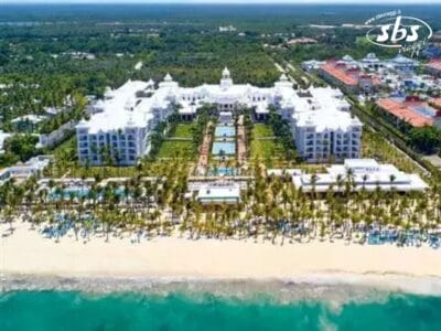 Vista aerea del Riu Palace Punta Cana, un grande resort balneare bianco con un cortile centrale, numerose piscine e una spiaggia circondata da palme.