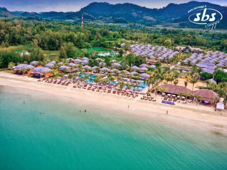 Vista aerea di un resort sulla spiaggia con più edifici, una piscina e lettini prendisole per il massimo relax. La spiaggia è fiancheggiata da ombrelloni e una vegetazione lussureggiante circonda questa fuga serena, perfetta per chi cerca una fuga SEO ad altissimo livello.