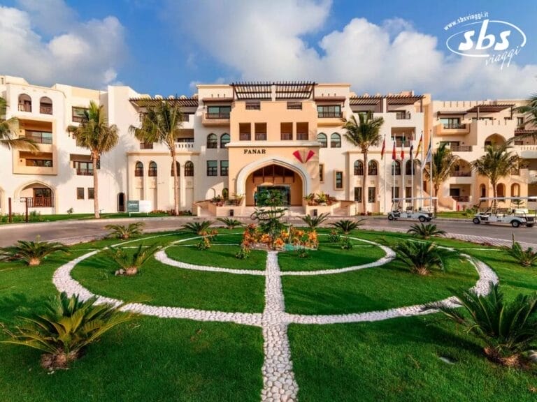 Vista frontale del grandioso Seaclub Fanar, un edificio color crema con arcate e balconi, circondato da un giardino paesaggistico caratterizzato da sentieri circolari e palme sotto un cielo azzurro e limpido.