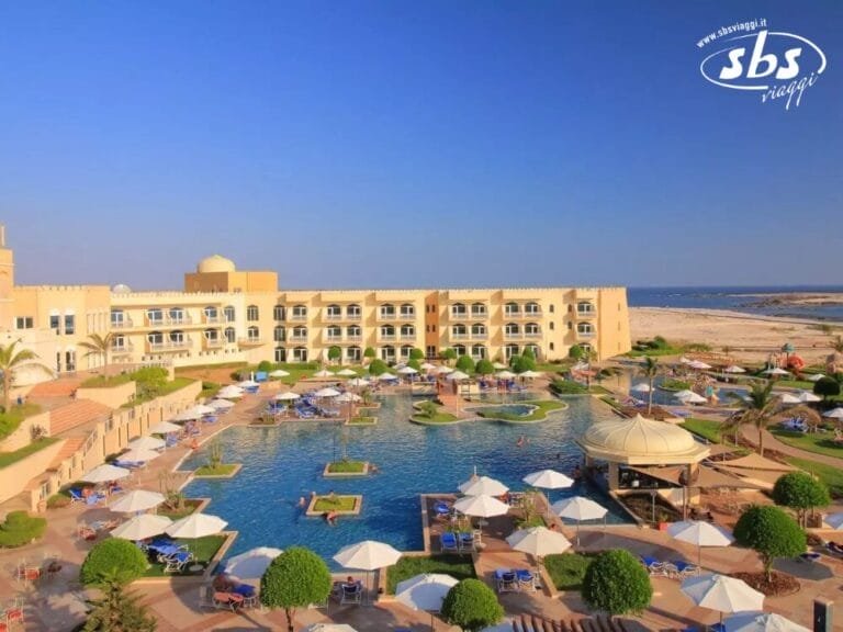 Vista aerea di un grande resort con piscina centrale, circondata da lettini e ombrelloni, che ricorda una bozza automatica di tranquillità, situato vicino alla costa sotto un cielo azzurro e limpido.