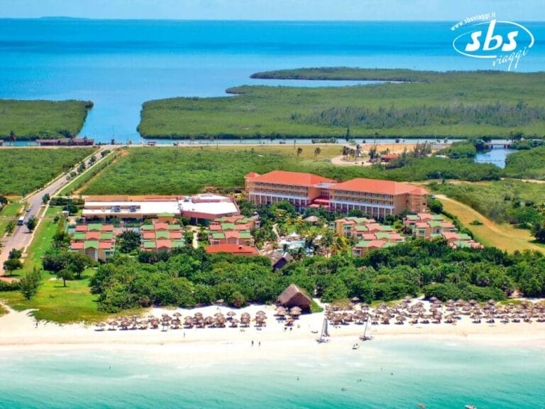 Vista aerea di un resort sulla spiaggia con l'oceano sullo sfondo, circondato da una vegetazione lussureggiante. Il resort Bozza automatica presenta diversi edifici e ombrelloni di paglia sparsi lungo la spiaggia incontaminata.