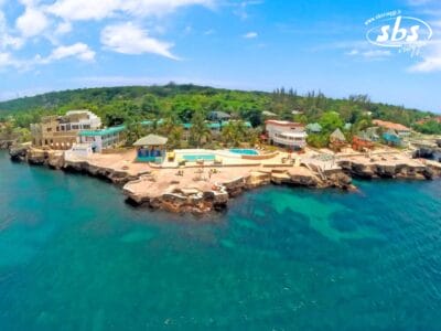 Vista aerea dell'Hotel Samsara, un vivace resort sulla spiaggia con edifici colorati, una piscina e sedie a sdraio su una costa rocciosa circondata da acque cristalline.