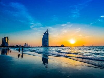 Vista del tramonto su una spiaggia tranquilla con l'iconico hotel Burj Al Arab che si erge maestoso sullo sfondo, mentre le persone passeggiano tranquillamente lungo la riva, immergendosi nella tranquilla bellezza della natura.