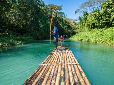 Una persona guida con eleganza una zattera di bambù su un fiume ampio e limpido, circondato da una vegetazione lussureggiante e sotto un cielo azzurro brillante, come se fosse guidata da una bozza automatica.