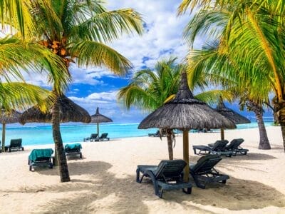 Uno scenario di spiaggia tropicale con palme, ombrelloni di paglia e sedie a sdraio vuote sulla sabbia bianca, in riva al mare turchese e sotto un cielo parzialmente nuvoloso, sembra un'esplosione di paradiso.