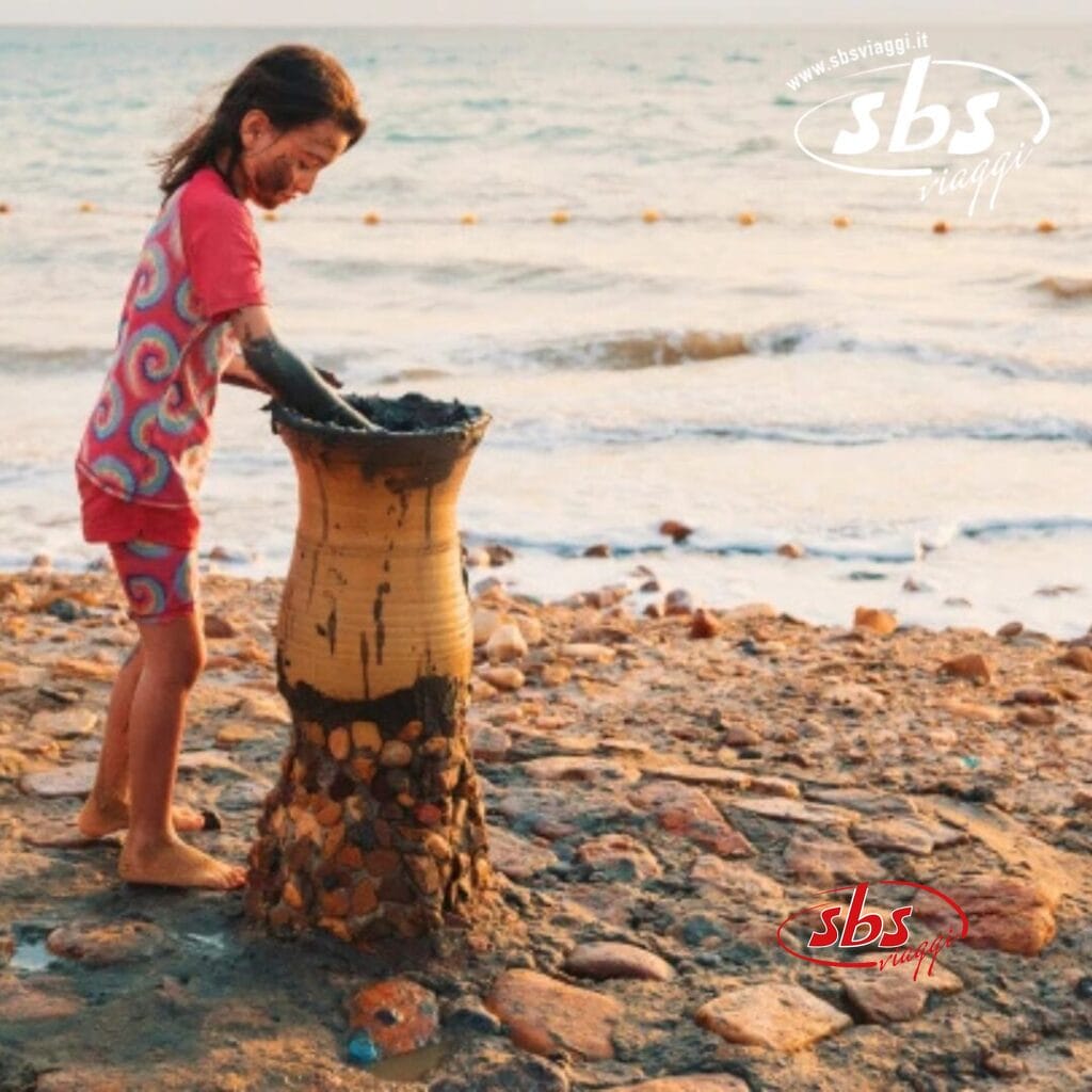 Un bambino in abiti colorati gioca con il fango su una spiaggia vicino a una struttura alta e ricoperta di fango, che ricorda gli incantevoli paesaggi della Giordania. L'oceano luccica sullo sfondo, trasformando la scena in un magico deserto in riva al mare.