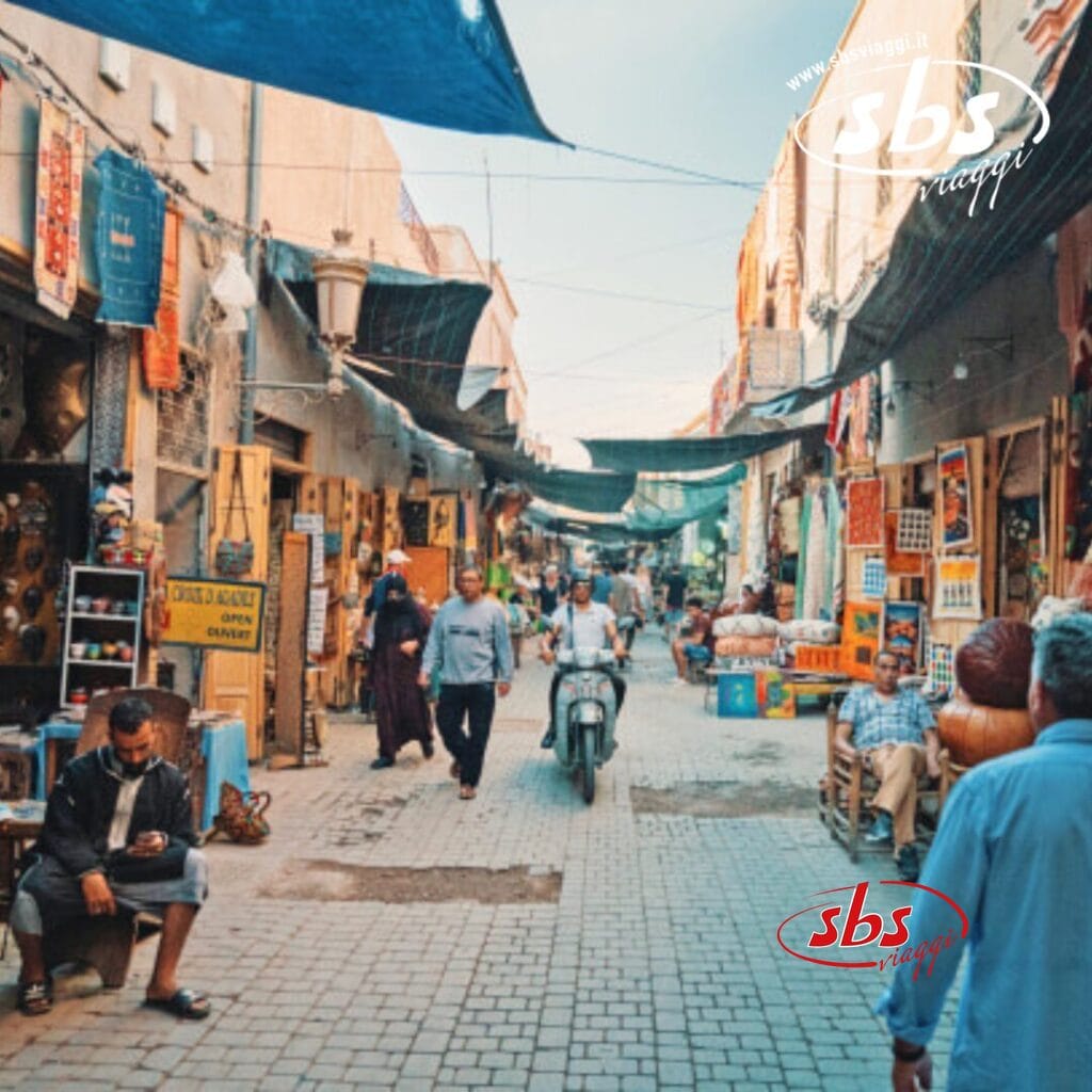 Un vivace mercato di strada a Marrakech con persone che camminano e una persona in motocicletta. I venditori espongono vari prodotti e tendoni colorati forniscono ombra. I cartelli sono visibili sui muri, creando un'atmosfera desertica vibrante in mezzo all'atmosfera vivace.