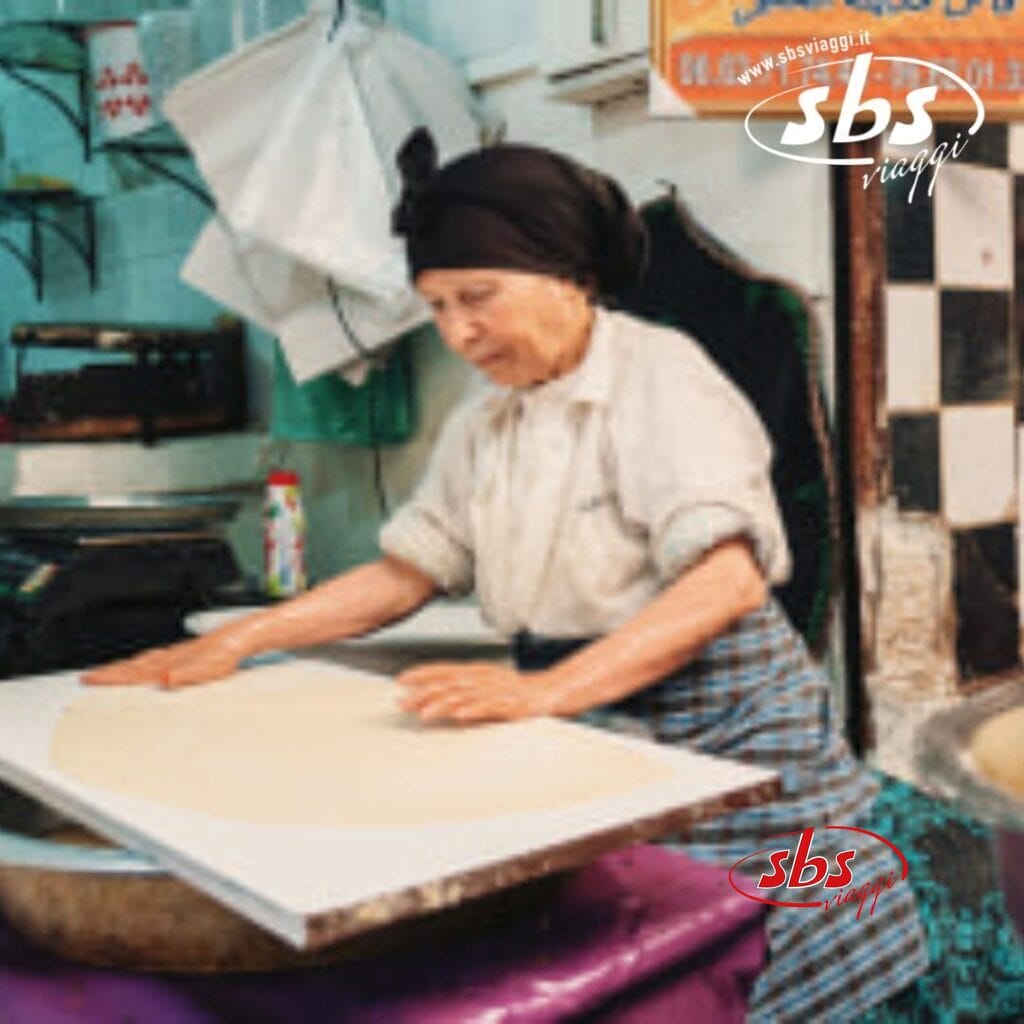 Una donna anziana con un foulard nero e una camicia leggera prepara l'impasto su una tavola di legno, che ricorda le scene tradizionali del Marocco. Utensili da cucina e una parete a scacchi fanno da sfondo a questo viaggio culinario.