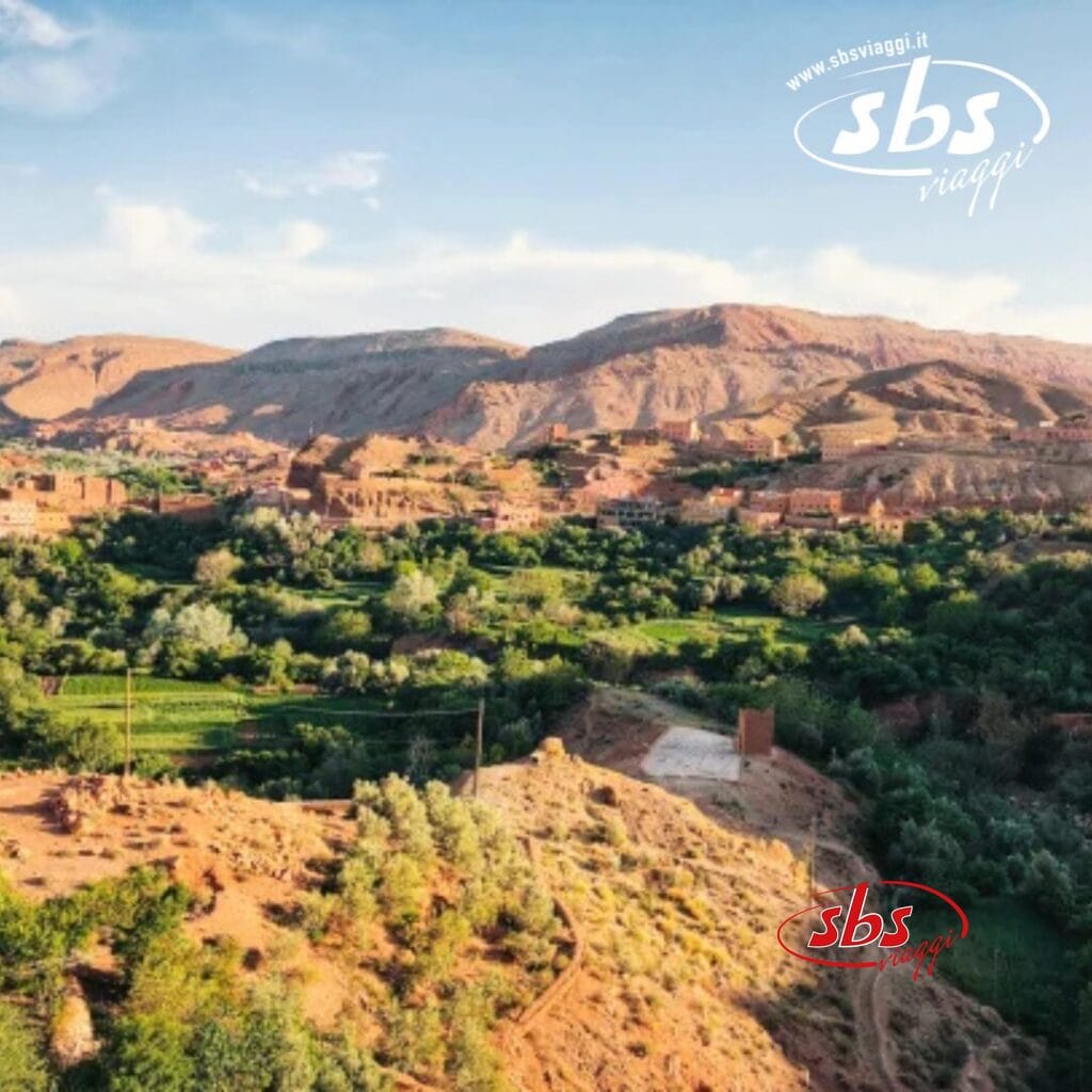 Vista aerea di una lussureggiante valle verde nel Grande Sud, circondata da montagne aride, con un piccolo insediamento e un'antica Kasbah visibili in lontananza sotto un limpido cielo azzurro del Marocco.