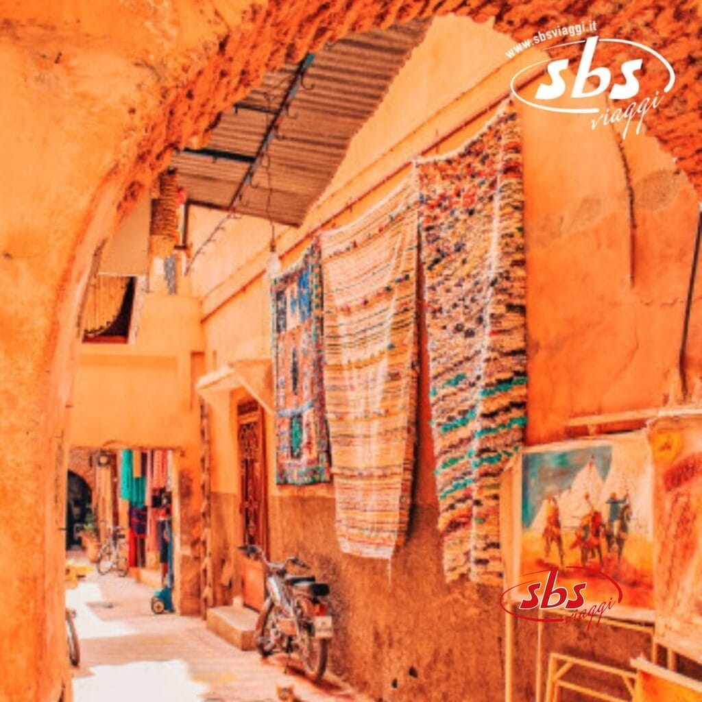 Un vicolo stretto e soleggiato che ricorda il Marocco, con tappeti colorati appesi a un muro e una moto parcheggiata. Sotto un arco, sono esposti tessuti squisiti e un dipinto, che catturano l'essenza dell'eleganza imperiale.