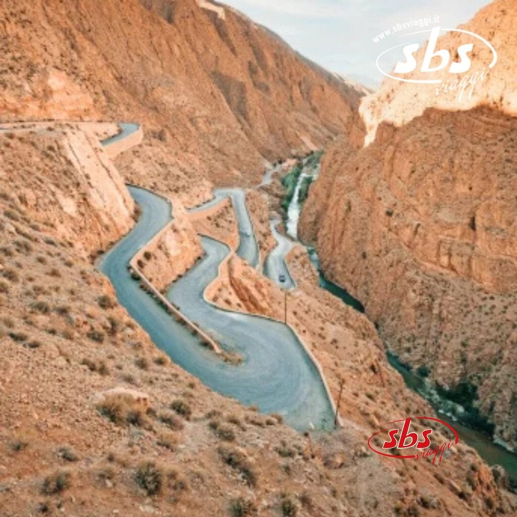 Una strada tortuosa con numerose curve strette scende attraverso il paesaggio roccioso e montuoso del Marocco, seguendo il percorso di un piccolo fiume sottostante.