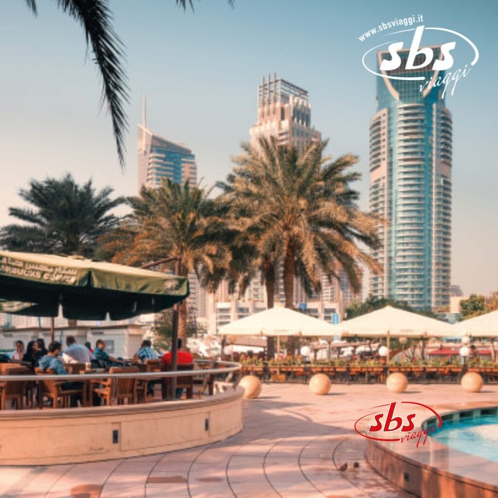 Bar all'aperto con clienti seduti sotto gli ombrelloni, palme e moderni grattacieli di Dubai sullo sfondo, con la filigrana 