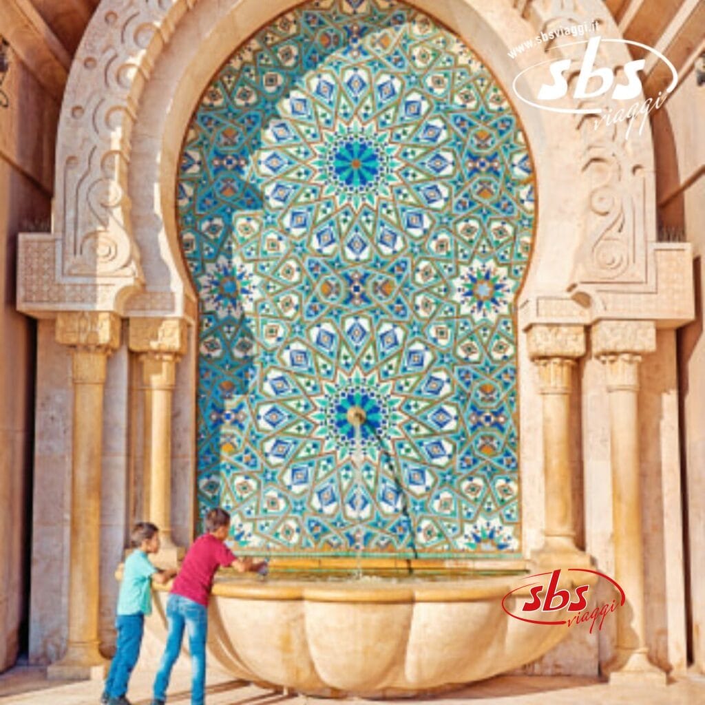 Due bambini sono in piedi accanto a una fontana decorativa a mosaico con un grande arco, decorato con intricati motivi blu e verdi che ricordano la vivace arte di Marrakech.