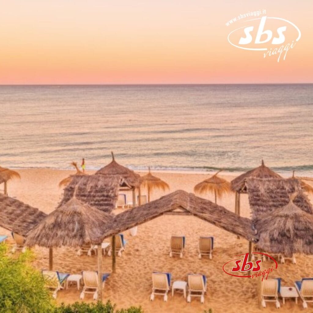 Scena di spiaggia al tramonto ad Hammamet, Tunisia, con ombrelloni di paglia e sedie a sdraio sulla sabbia. Le onde calme nell'oceano riflettono un cielo caldo, arancione-rosa, che si fonde perfettamente con la ricca storia della regione.