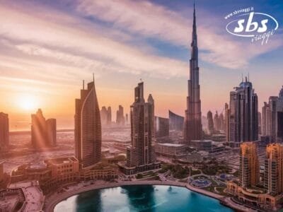 Una vista aerea dello skyline della moderna città di Dubai al tramonto rivela imponenti grattacieli e uno specchio d'acqua scintillante in primo piano. Il cielo parzialmente nuvoloso è dipinto in vivaci tonalità di arancione e viola.