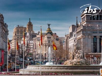 Piazza della città di Madrid con la fontana di Cibele in primo piano, circondata da edifici storici e bandiere spagnole, sotto un cielo azzurro e terso che ricorda una perfetta giornata di giugno.