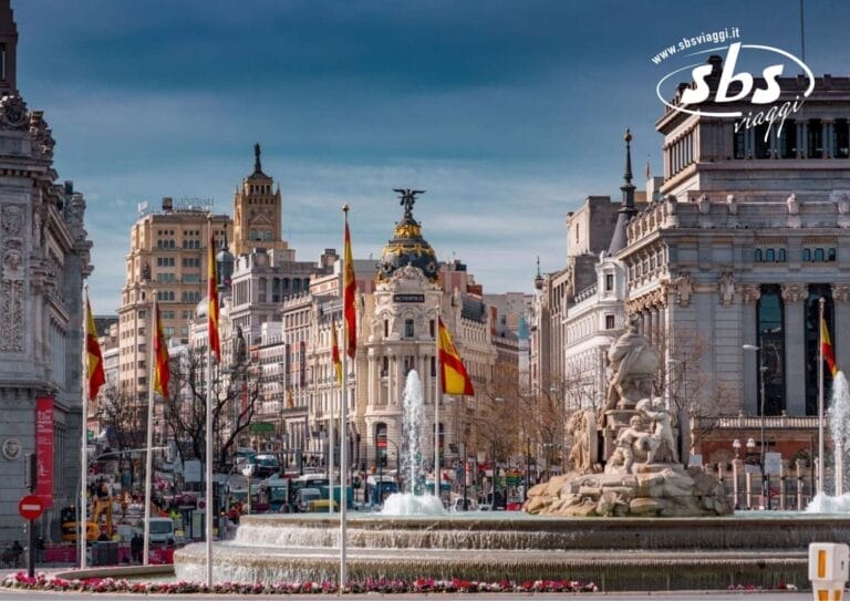 Piazza della città di Madrid con la fontana di Cibele in primo piano, circondata da edifici storici e bandiere spagnole, sotto un cielo azzurro e terso che ricorda una perfetta giornata di giugno.