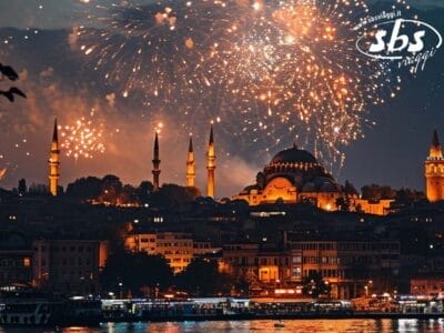 I fuochi d'artificio illuminano il cielo notturno sopra un paesaggio urbano con una moschea e minareti splendidamente illuminati, i cui colori si riflettono sull'acqua sottostante in una scena che potrebbe fungere da bozza automatica per immagini mozzafiato.