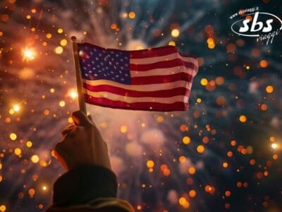 Una mano regge una piccola bandiera americana sullo sfondo dei fuochi d'artificio di Capodanno e delle luci brillanti di una città americana.