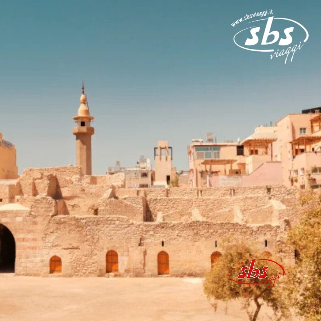 Edifici storici in pietra con ingressi ad arco, un minareto che si erge sotto il cielo terso e azzurro, evocano l'incantevole fascino della Giordania. Nell'angolo in alto a destra, sono visibili i loghi 