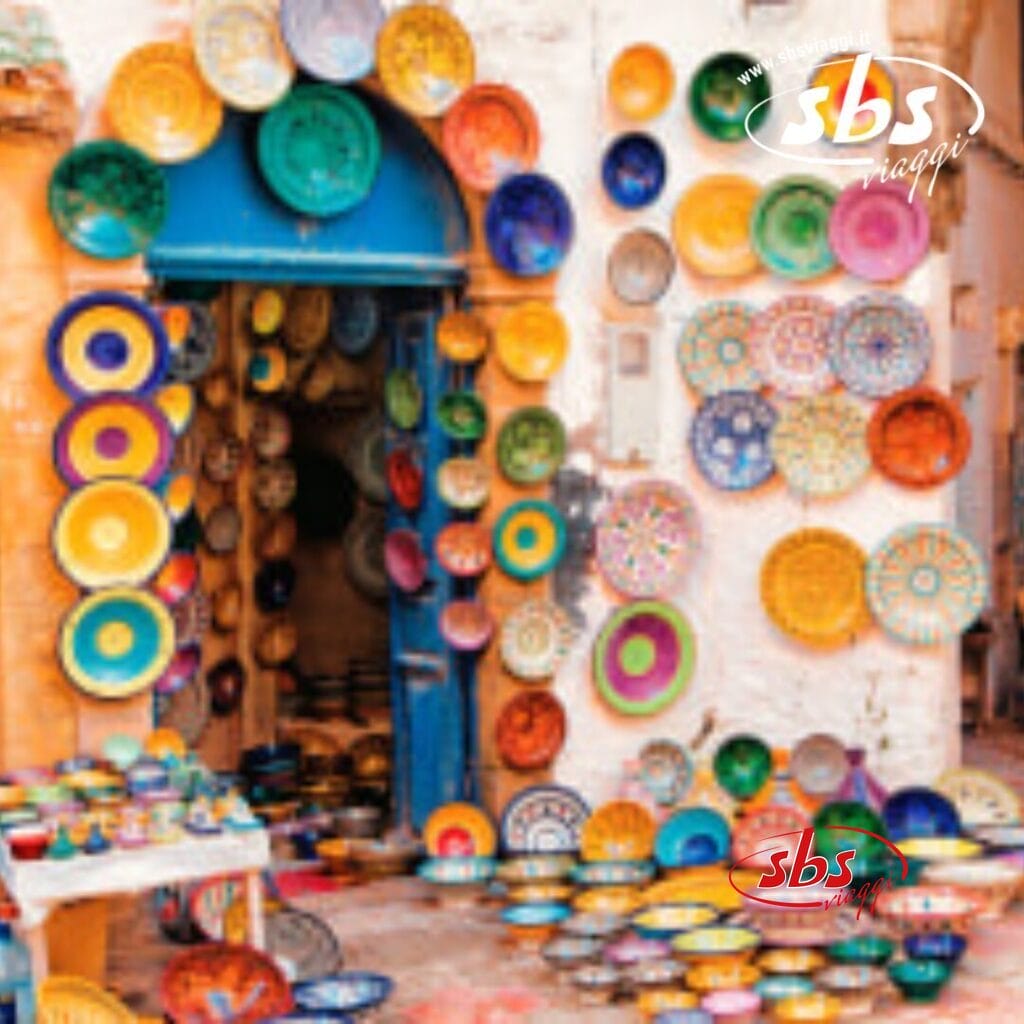 Piatti di ceramica colorati, che ricordano i mercati di Marrakech, sono esposti su una parete e per terra, all'esterno di un negozio con una porta blu.