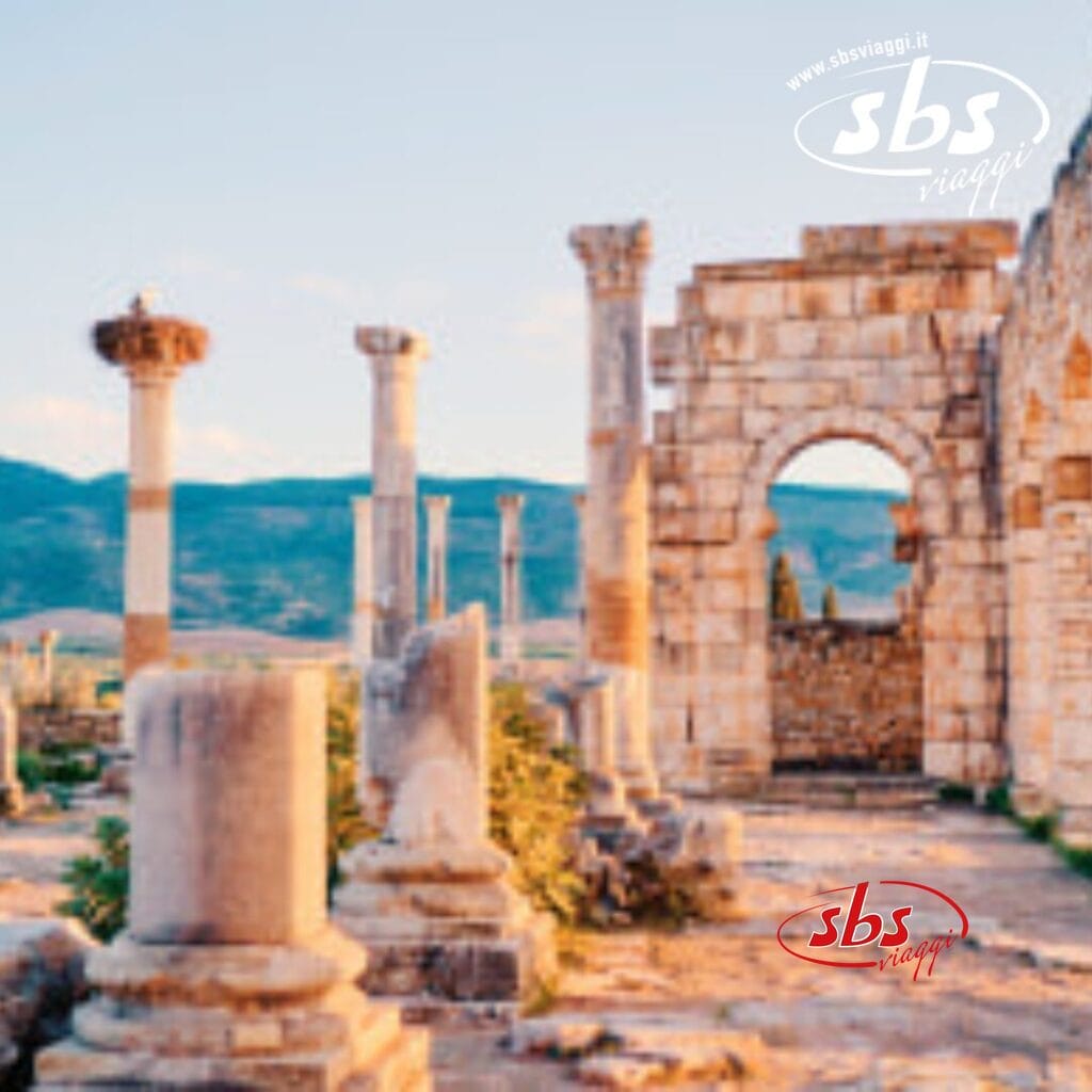 Le rovine di antiche colonne di pietra e un arco si ergono fiere sullo sfondo di montagne e cielo limpido, parte degli incantevoli panorami del Gran Tour del Marocco, come descritto in dettaglio nel Programma 2024.