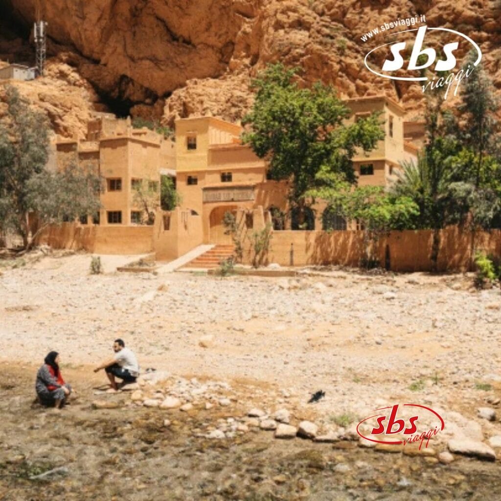 Due persone sono sedute vicino a un ruscello roccioso, con sullo sfondo rustici edifici desertici che ricordano una Kasbah e le aspre scogliere del Grande Sud del Marocco, il tutto sotto un cielo limpido.