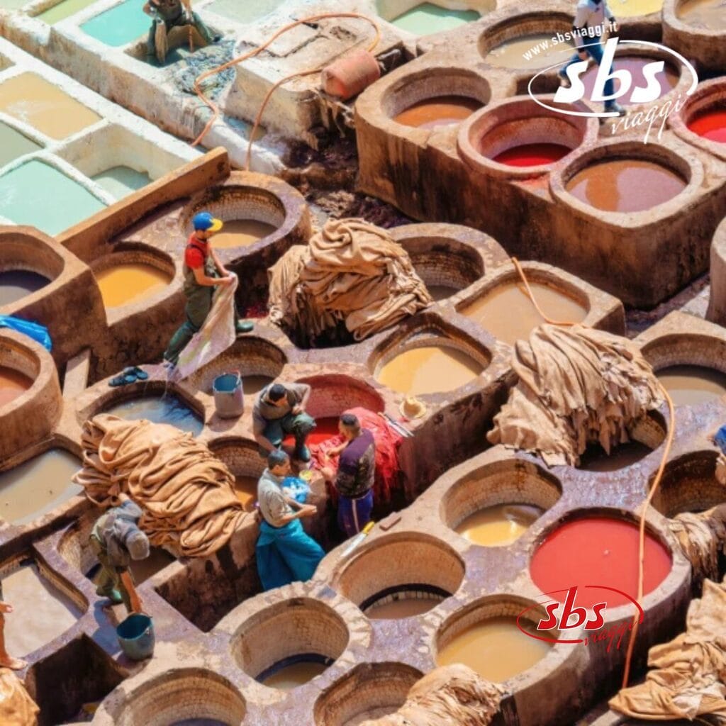 I lavoratori della tradizionale conceria di pelli Marocco Imperiale sono visti dall'alto, mentre lavorano duramente su grandi tini rotondi pieni di liquidi colorati e pelli. L'antico mestiere si dispiega sotto di loro, un vivido arazzo di toni intensi e arte senza tempo.