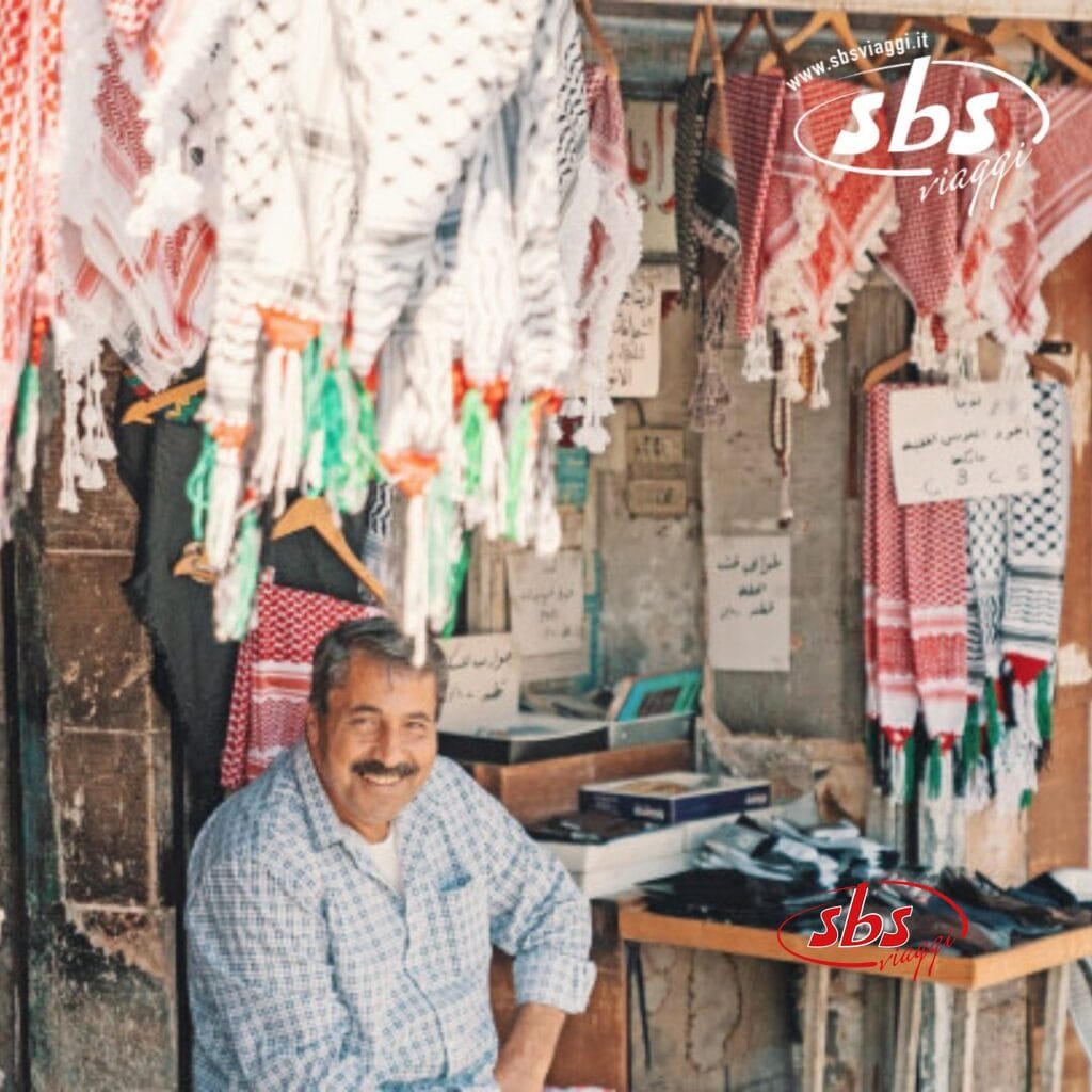 Un uomo è seduto in un negozio in Giordania, dove vende sciarpe tradizionali con scritte in arabo sui cartelli, invitando i turisti a scoprire le meraviglie dell'artigianato locale.