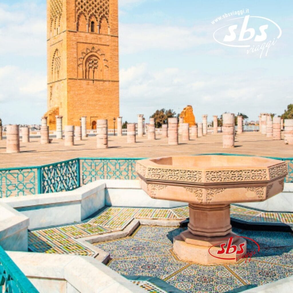 Una fontana geometrica in pietra in un cortile piastrellato con alte colonne e una torre che ricorda la medine del Nord in Marocco. Ringhiere blu circondano l'area, aggiungendone fascino.