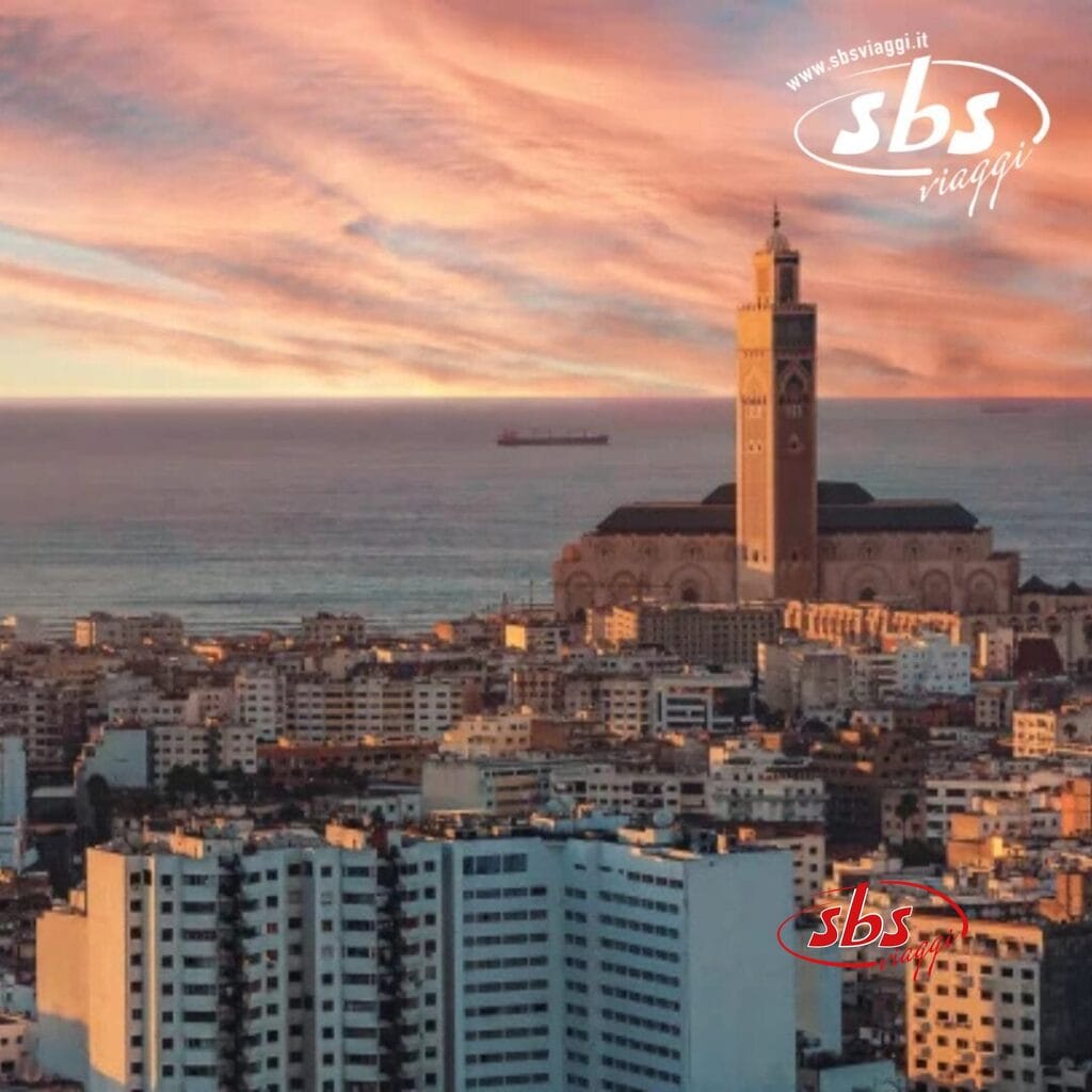 Paesaggio urbano di Casablanca con edifici imponenti, l'iconica moschea di Hassan II sull'oceano e un vivace cielo al tramonto, che catturano perfettamente il fascino del Marocco.