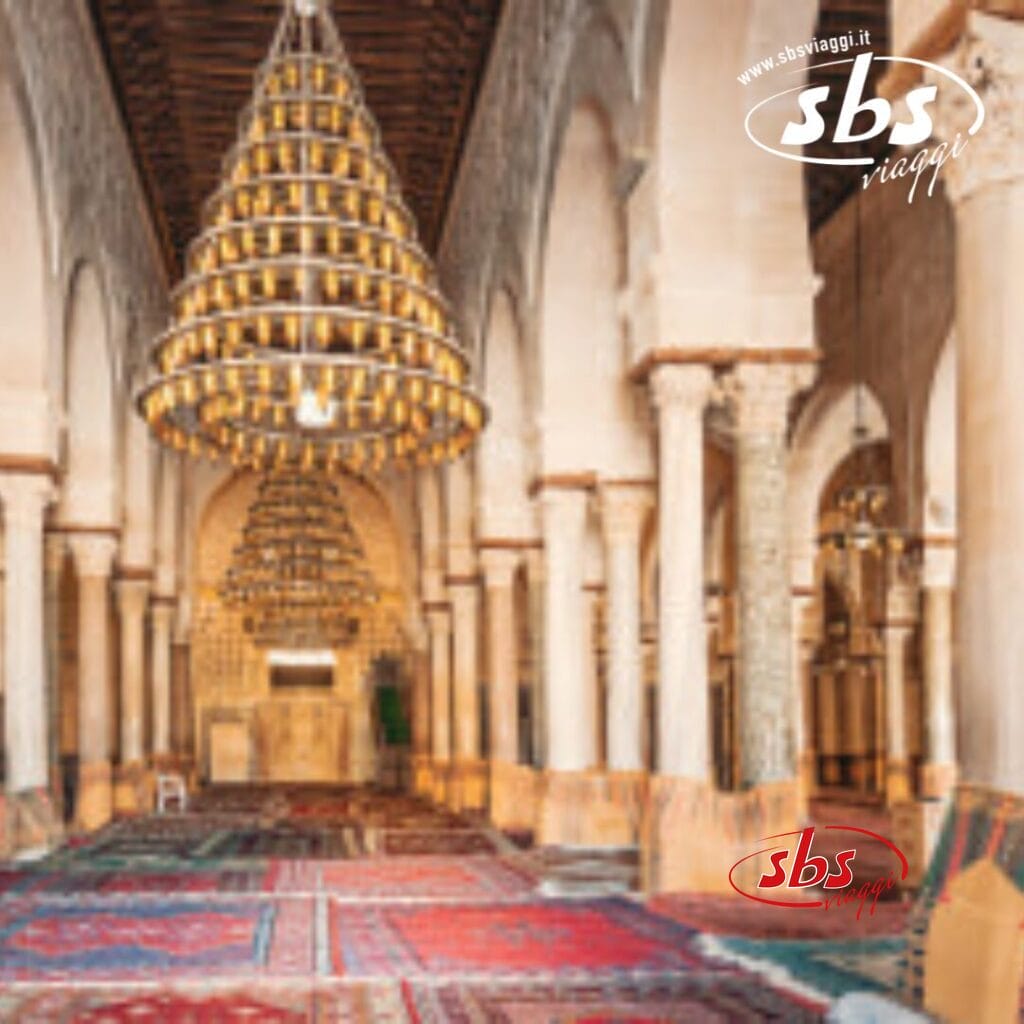 L'interno della moschea di Hammamet, Tunisia, presenta un soffitto alto decorato con lampadari decorati e una fila di colonne. Il pavimento è splendidamente ricoperto di tappeti colorati, creando un'atmosfera che riflette il ricco patrimonio culturale di questo luogo storico.