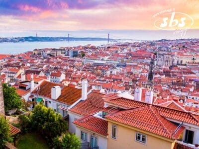 Una vista panoramica di Lisbona con i tetti rossi, il Ponte 25 de Abril e il fiume Tago sullo sfondo di un cielo colorato al tramonto.