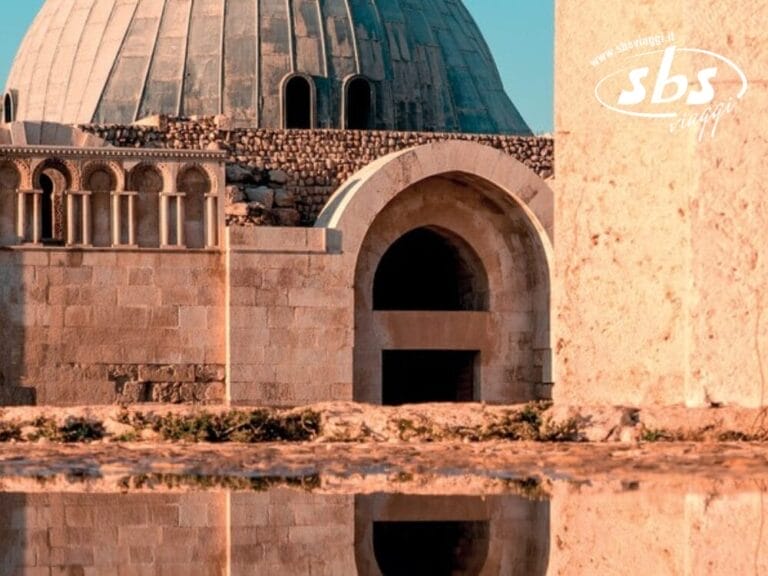 Edificio in pietra con un grande tetto a cupola e ingressi ad arco, riflesso in uno specchio d'acqua. Il logo nell'angolo recita 