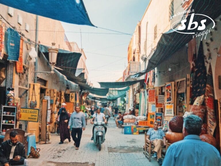 Una strada di mercato movimentata, che ricorda una vivace Kasbah, vede persone che si muovono tra la folla vivace e una persona che scivola su uno scooter. I negozi sotto le tettoie espongono una serie di merci, riecheggiando la ricca cultura e lo spirito del Grande Sud del Marocco.